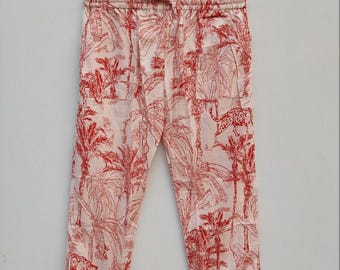 May include: Light pink pants with a red tropical print featuring palm trees and leopards. The pants have a drawstring waist and are hanging on a wooden hanger.