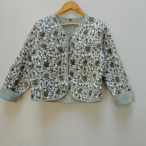 May include: A white jacket with a green floral pattern and green striped trim. The jacket has long sleeves and a V-neck. It is hanging on a wooden hanger against a plain background.