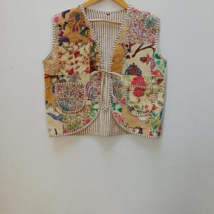 May include: A cream-colored quilted vest with a tie closure and floral patchwork design. The vest has two pockets on the front and a striped lining.