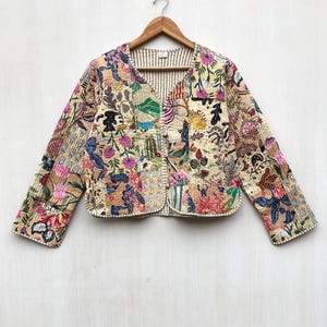 Puede incluir: Chaqueta acolchada beige con cuello en V, adornada con un patchwork de diseños florales y abstractos en rosa, azul y verde. La chaqueta, colgada de una percha de madera, muestra un frente abierto y costuras detalladas.