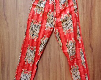 May include: Red pajama pants with vertical stripes and a repeating tiger print. The pants are hanging on a wooden hanger against a wooden background. The tiger print features black and tan stripes.