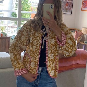 May include: Mustard yellow quilted jacket with a white floral design. The jacket has red and white striped trim on the cuffs and pockets. It features a drawstring closure and is paired with blue jeans.
