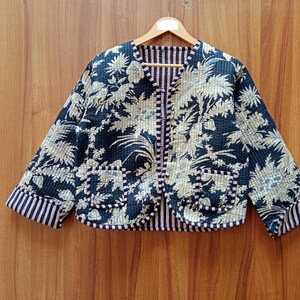 May include: A quilted jacket with a floral pattern in navy blue and beige. The jacket has a striped lining and trim, with two front pockets. It is displayed on a wooden hanger against a wooden background.