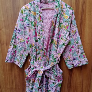 May include: A pink floral patterned robe with a matching belt. The robe features a quilted design and is adorned with green, yellow, and purple floral patterns. The robe is hanging on a wooden hanger.