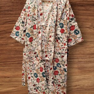 May include: A cream-colored floral robe with a white trim, hanging on a wooden hanger. The robe features a vibrant pattern of red, blue, and yellow flowers and green leaves. The robe has a tie belt and short sleeves.