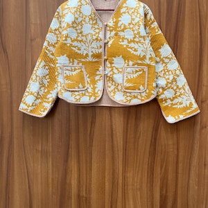 May include: A mustard yellow quilted jacket with a white floral print. The jacket has a button closure and two pockets.
