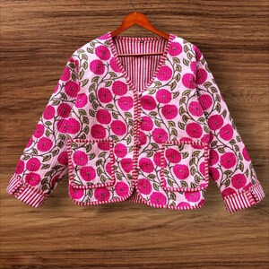 May include: A quilted jacket with a floral pattern in shades of pink and white. The jacket has a V-neck, two pockets, and red and white striped cuffs. The jacket is hanging on a wooden hanger.