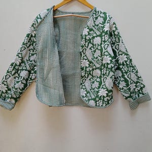 Könnte beinhalten: Eine reversible Jacke in Grün und Weiß mit einem Blumendruck auf der einen Seite und einem gestreiften Muster auf der anderen. Die Jacke hängt an einem Holzbügel.