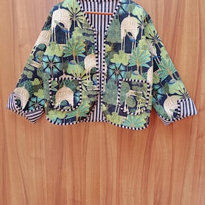 May include: A quilted jacket with a botanical print of trees and birdcages in green, blue, and beige. It has a black and white striped lining and trim, two front pockets, and a V-neck. The jacket is hung on a wooden hanger.