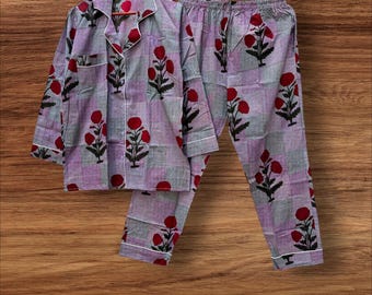 May include: Lavender and grey checked pyjama set with red floral patterns. The long-sleeved top and trousers have white trim. The set is displayed on wooden hangers against a wood grain background. The pyjamas are made of a soft material.