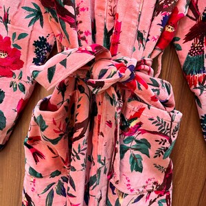 Peut inclure: Une robe en velours rose avec un imprim&eacute; floral repr&eacute;sentant des oiseaux et des feuilles. La robe a une ceinture &agrave; nouer et deux poches.