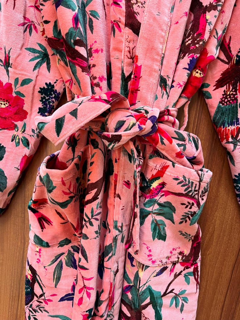 Peut inclure: Une robe en velours rose avec un imprim&eacute; floral repr&eacute;sentant des oiseaux et des feuilles. La robe a une ceinture &agrave; nouer et deux poches.