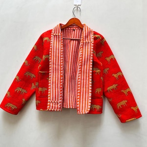 May include: A red quilted jacket with a tiger print. The jacket has long sleeves and a collared neckline. The interior lining features orange and white vertical stripes. The jacket is hanging on a wooden hanger.