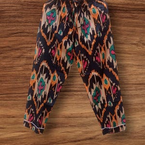 Peut inclure: Un pantalon de pyjama à motifs avec une taille à cordon de serrage, suspendu à une tringle en bois. Le pantalon présente un fond bleu marine avec un motif de style ikat en orange, sarcelle et rose. Les poignets ont une bordure blanche.