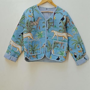 Puede incluir: Chaqueta acolchada azul claro con estampado de animales de la jungla con cebras, leopardos y tucanes. La chaqueta tiene cuello en V, dos bolsillos y ribete a rayas en los puños y bordes. La chaqueta está colgada en una percha de madera.