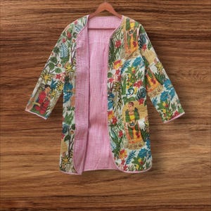 May include: A long, open-front jacket with a colorful floral and figure print on a white background. The jacket's interior is a light pink with vertical stripes. The jacket is hanging on a wooden hanger against a wooden background.