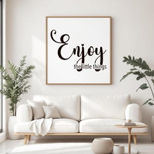 Könnte beinhalten: Ein gerahmter Wandkunstdruck mit dem Schriftzug "Enjoy the little things" in schwarzer Schrift. Das Kunstwerk wird über einem weißen Sofa mit dekorativen Kissen präsentiert. Der Raum ist mit Pflanzen und einem kleinen Beistelltisch aus Holz dekoriert.