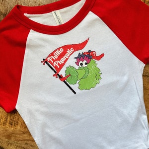 May include: White and red baseball style t-shirt with a graphic of the Phillie Phanatic mascot holding a banner that says "Phillie Phanatic". The shirt is made of a soft material.