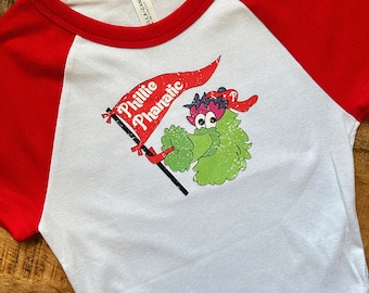 May include: White and red baseball style t-shirt with a graphic of the Phillie Phanatic mascot holding a banner that says "Phillie Phanatic". The shirt is made of a soft material.
