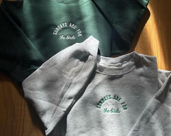 May include: Two sweatshirts, one forest green and one gray, are displayed on a wooden surface. Each sweatshirt has the embroidered text "SUNDAYS ARE FOR the birds" in a semi-circle. The green sweatshirt is on top, with the gray one below.