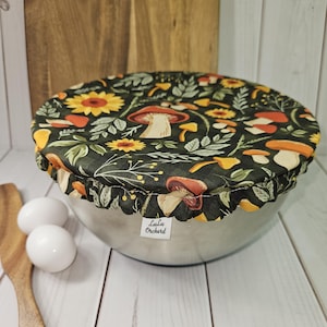 May include: A round fabric bowl cover with an elastic edge, featuring a dark green background with a pattern of yellow sunflowers, various mushrooms, and green leaves. A small white tag with the text LuLu Orchard is attached to the side.