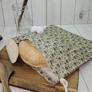 May include: A loaf of bread partially inside a floral patterned bread bag, with a slice of bread on a wooden cutting board. Two white eggs and a glass jar with a wooden spoon are in the background. The bag has a drawstring closure.