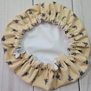 May include: A circular bowl cover featuring a light tan fabric exterior with a repeating pattern of small black bees. The cover has an elasticized gathered edge and a solid white interior lining, designed to fit snugly over the rim of a container.