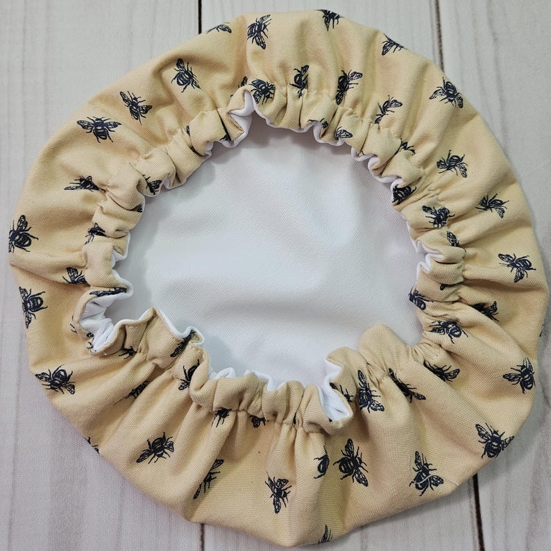 May include: A circular bowl cover featuring a light tan fabric exterior with a repeating pattern of small black bees. The cover has an elasticized gathered edge and a solid white interior lining, designed to fit snugly over the rim of a container.