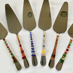 May include: Seven silver cake servers with beaded handles. The handles are decorated with colourful beads in various patterns, including red, orange, yellow, green, blue and purple.