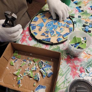 May include: A mosaic art project in progress. A wooden circle is partially covered with blue and green ceramic tile shards. A person wearing gloves is working on the mosaic, with tools and additional tile pieces nearby. The project is on a floral patterned surface.