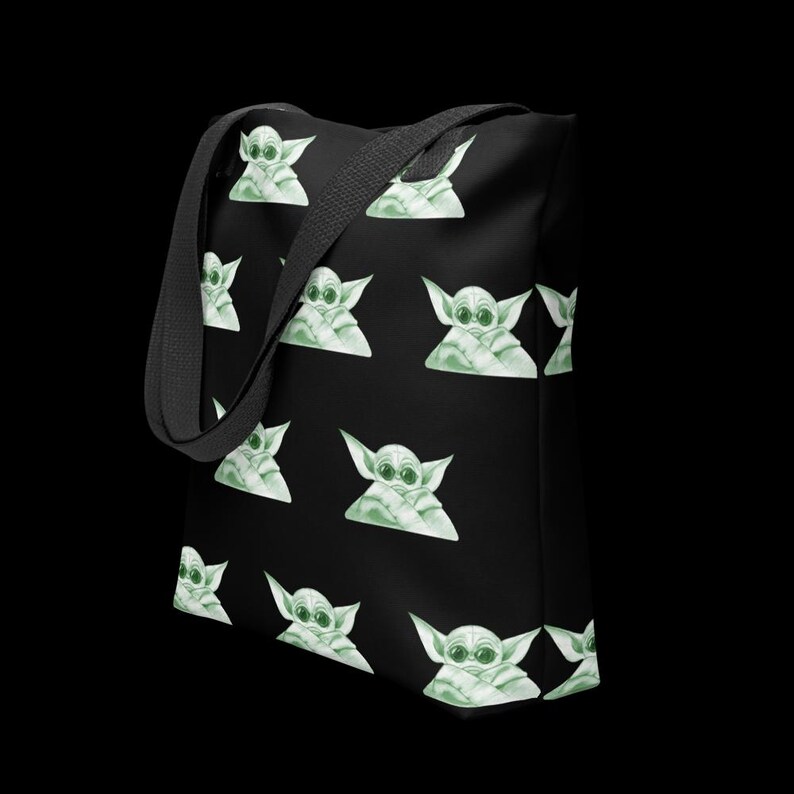 May include: Black tote bag featuring a repeating pattern of a green and white cartoon character. The bag has a black shoulder strap and is set against a black background. The cartoon character has large eyes and pointed ears.