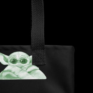 May include: Black tote bag featuring a green illustration of a character with large eyes and pointed ears. The bag has a black strap and is made of a durable material. The design is a popular character from a science fiction franchise.