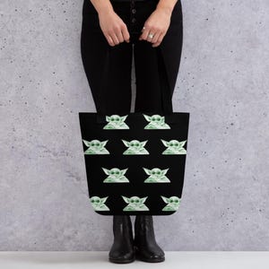 May include: Black tote bag featuring a repeating pattern of a green alien character with large ears and sunglasses. The bag is held by a person wearing black pants and boots. The background is a plain gray wall.
