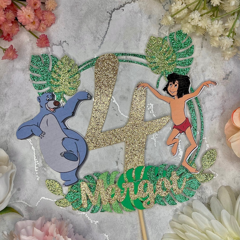 May include: A gold glitter number four cake topper with green glitter leaves and a jungle theme. The topper features a cartoon image of Mowgli from The Jungle Book, Baloo the bear, and the text "Mowgli".