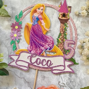 May include: A purple and gold glitter cake topper featuring Rapunzel from the Disney movie Tangled. The topper includes a banner with the name "Coco" and a tower with vines.