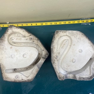 以下が含まれることがあります： 白く塗られた2つの白鳥の形をした石膏型。型は少し風化していて、ざらざらした質感です。