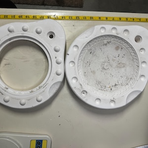 Puede incluir: Dos objetos circulares blancos con una serie de protuberancias elevadas alrededor del borde exterior. Uno tiene una abertura central, el otro un interior empotrado. Una cinta métrica amarilla indica unos 25 cm de diámetro.