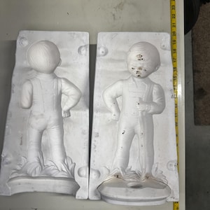 May include: Two-part plaster mold of a young boy in overalls, standing with hands on hips. The white mold shows the boy's form in detail, with visible facial features and clothing folds. The mold is on a light-colored surface.