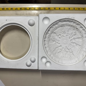 Puede incluir: Molde de yeso blanco de dos partes. Una sección tiene una cavidad circular, la otra presenta un diseño en relieve detallado. El molde es cuadrado con esferas redondeadas en las esquinas. Una cinta métrica amarilla está en la parte superior.