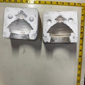 May include: Two white molds with a decorative glass object inside. The glass object has a tiered, pagoda-like shape. A yellow measuring tape is visible along the top and right side of the image.