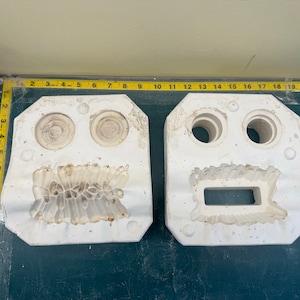 May include: Two white plaster molds for casting. The mold on the left has two round impressions and a detailed, textured design. The mold on the right has two round impressions and a rectangular impression.