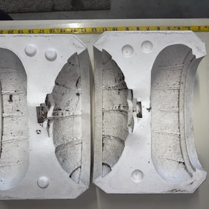 Può includere: Stampo bianco in due pezzi con un design curvo, probabilmente per la fusione. Lo stampo è aperto, rivelando la forma interna. Un metro a nastro giallo è posizionato lungo il bordo superiore, con misure in centimetri.