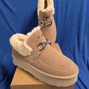May include: A pair of khaki platform slip-on shoes with cream-colored faux fur lining and a silver-toned metal chain detail across the top. The shoes feature the UGG logo on the front and sit on a brown cardboard box labeled ART:A25, COLOR:KHAKI, SIZE:9.