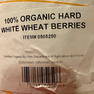 May include: A white label with black text that reads "100% ORGANIC HARD WHITE WHEAT BERRIES" and "ITEM# 0505250". The label also includes the text "Certified Organic by Utah Department of Agriculture and Food" and "CONTAINS ORGANIC WHEAT". A black and white barcode is printed below the text. The barcode number is "0 75211 05250 6".