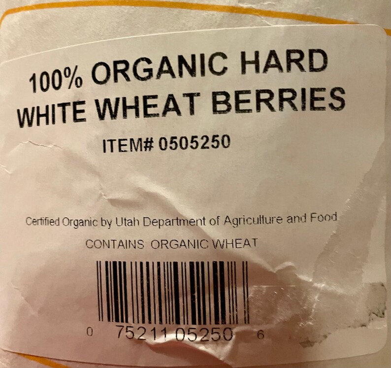 May include: A white label with black text that reads "100% ORGANIC HARD WHITE WHEAT BERRIES" and "ITEM# 0505250". The label also includes the text "Certified Organic by Utah Department of Agriculture and Food" and "CONTAINS ORGANIC WHEAT". A black and white barcode is printed below the text. The barcode number is "0 75211 05250 6".