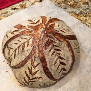 May include: A freshly baked loaf of sourdough bread with a decorative leaf pattern scored into the top crust. The bread is golden brown and has a rustic appearance.