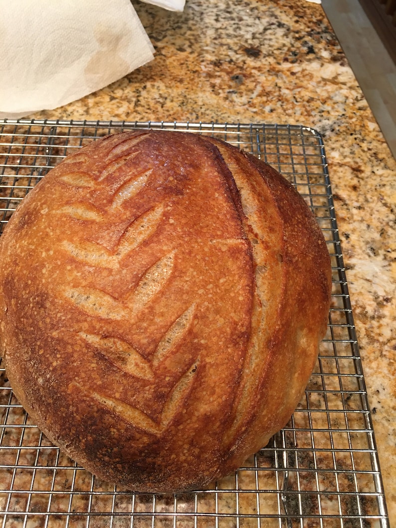 May include: A freshly baked loaf of sourdough bread with a deep brown crust and a decorative leaf pattern scored into the top.