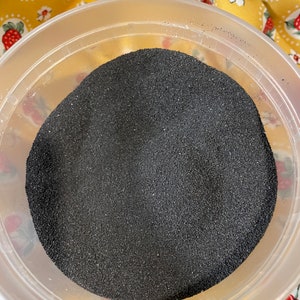 K&ouml;nnte beinhalten: Ein durchsichtiger Plastikbeh&auml;lter gef&uuml;llt mit schwarzem Sand. Der Beh&auml;lter steht auf einem gelben und wei&szlig;en Stoff mit einem Kirschmuster.