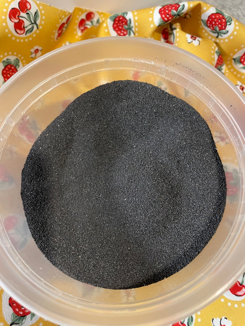 K&ouml;nnte beinhalten: Ein durchsichtiger Plastikbeh&auml;lter gef&uuml;llt mit schwarzem Sand. Der Beh&auml;lter steht auf einem gelben und wei&szlig;en Stoff mit einem Kirschmuster.