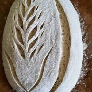 May include: A close-up of a loaf of sourdough bread with a wheat design scored into the top. The bread is dusted with flour and has a light brown crust.
